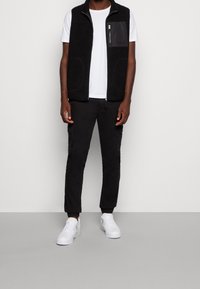 Muž v černé fleecové vestě přes bílé tričko s krátkým rukávem, černých cargo kalhotách a bílých teniskách stojí na jednobarevném pozadí.