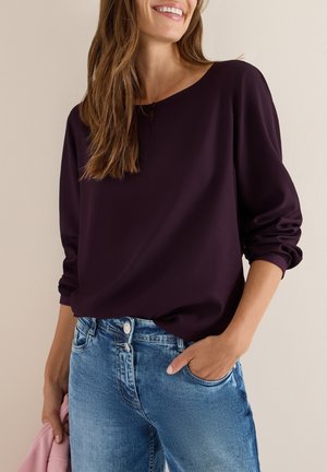 Femme portant un haut violet foncé à manches longues et un jean bleu, une main dans la poche, tenant un tissu rose dans l'autre main.