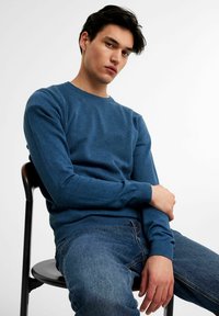 Blauer Strickpullover mit Rundhalsausschnitt und langen Ärmeln, kombiniert mit Jeans. Der Pullover hat gerippte Bündchen und Saum.