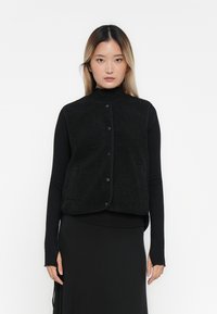 Zwarte fleece vest met een hoge kraag, drukknoopsluiting en twee voorzakken. Draag over een zwart longsleeve shirt en rok. Textuur oppervlak.