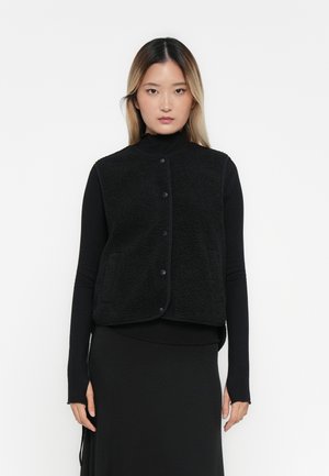 Gilet di pile nero con colletto alto, bottoni a pressione e due tasche frontali. Indossato sopra una maglia nera a maniche lunghe e una gonna. Superficie testurizzata.