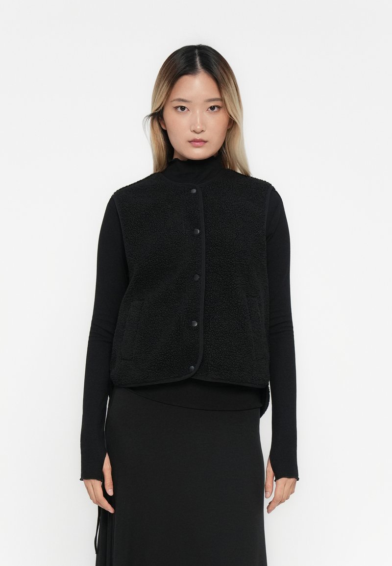 Zwarte fleece vest met een hoge kraag, drukknoopsluiting en twee voorzakken. Draag over een zwart longsleeve shirt en rok. Textuur oppervlak.