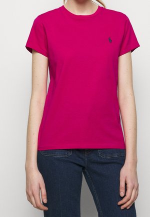 Kobieta ubrana w prosty, jasnoróżowy t-shirt z krótkim rękawem, zestawiony z ciemnoniebieskimi jeansami na jednolitym tle.