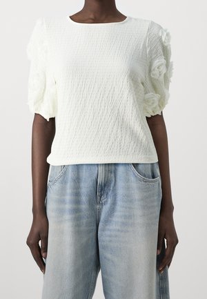 Kvinde iført tekstureret hvid kortærmet top med 3D-blomsterdetaljer på ærmerne og lyseblå højtaljede jeans.