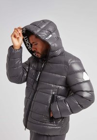 Chaqueta acolchada gris con capucha, con diseño acolchado horizontal, dos cremalleras frontales y un logotipo en el interior y en la manga izquierda.