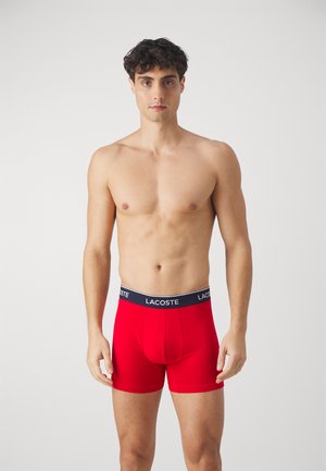 Boxers rojos hechos de tela elástica con una cintura de la marca Lacoste en negro. Presentan un ajuste ceñido y una longitud hasta medio muslo.