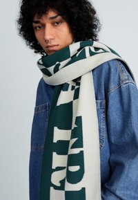 Jovem com cabelo encaracolado a usar um lenço verde e branco com padrão e uma jaqueta de ganga azul, diante de um fundo simples.