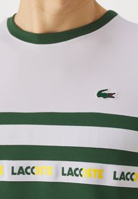 Bílá a zelená pruhovaná košile Lacoste se zeleným límečkem. Má texturovanou tkaninu a logo Lacoste v zelené a červené barvě na hrudi.