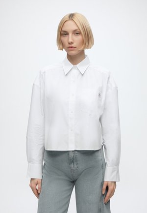 Jeune femme aux cheveux blonds courts portant une chemise courte blanche boutonnée et un jean taille haute bleu clair, debout devant un fond uni.