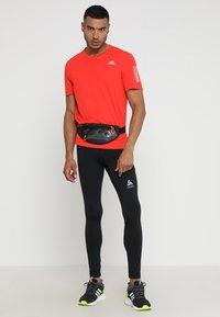 Rotes kurzärmliges Sportshirt, schwarze eng anliegende Leggings und eine schwarze Hüfttasche mit orangefarbenen Akzenten, getragen mit schwarzen und neongelben Laufschuhen.