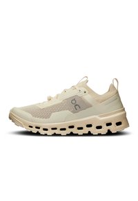 Chaussure de sport beige avec une tige en mesh texturé, une semelle amortie et des découpes décoratives. Elle est dotée de lacets et d'une languette de talon renforcée.