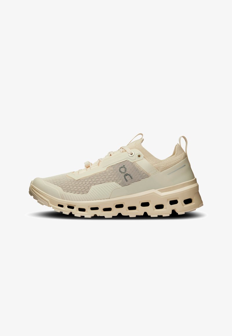 Chaussure de sport beige avec une tige en mesh texturé, une semelle amortie et des découpes décoratives. Elle est dotée de lacets et d'une languette de talon renforcée.