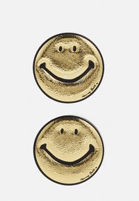 Pins de sorriso dourados metálicos com uma superfície refletora, apresentando um design de sorriso simples e uma borda preta. Assinatura na borda inferior.