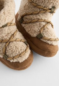 Beige Kunstfellstiefel mit braunen Wildledersohlen, ausgestattet mit kreuzenden Schnürsenkeln in einem tan- und braunfarbenen Muster. Weicher Stoff, runde Form und Knöchelhöhe.