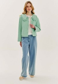 Světlonzelená prošívaná bunda s velkým límcem, předními knoflíky a dvěma kapsami, v kombinaci s volnými modrými džíny a béžovými botami.