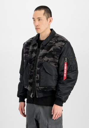 Mann trägt schwarze und graue Fleecemantel mit Camouflage-Muster, großen Taschen und einem roten "Remove Before Flight"-Etikett an der Ärmeltasche.