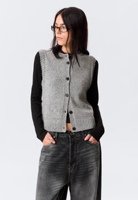 Femme aux longs cheveux bruns et portant des lunettes, vêtue d'un gilet en tricot gris à boutons sur un t-shirt noir à manches longues et un jean large foncé, les mains dans les poches.