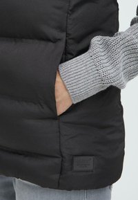 Un gros plan de la main d'une personne dans une poche d'un gilet en doudoune noir, avec une manche de pull en tricot gris visible. Logo de la marque affiché.