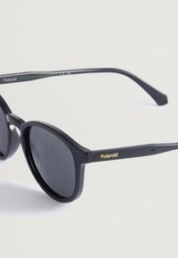 Μαύρα πλαστικά γυαλιά ηλίου με στρογγυλούς φακούς, με το χρυσό λογότυπο "Polaroid" στο βραχίονα. Φακοί με γκρίζους τόνους, κομψός σχεδιασμός.