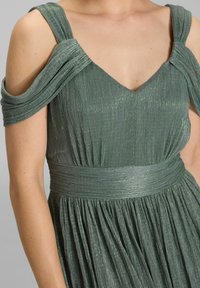 Robe plissée verte avec un décolleté en V profond, des bretelles dénudées et un tissu texturé. Présente une taille cintrée avec une large ceinture.