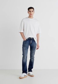 White oversized t-shirt, dark blue jeans with a slight fade, and white sneakers with black accents and gum soles. Standing pose on a plain background.