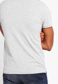 Persoon in een lichtgrijs T-shirt en donkerblauwe jeans, met de armen achter de rug gekruist, gefotografeerd van achteren op een witte achtergrond.