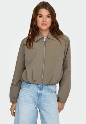 Mladá žena v taupe mikine na zips a svetlomodrých džínsach s vysokým pásom, stojaca pred jednoduchým bielym pozadím.