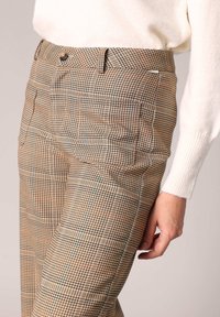 Pantalons à carreaux dans des tons marron avec un motif subtil. Présentent un devant plat, des poches latérales et une fermeture à bouton. Le matériau semble texturé.