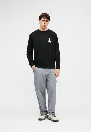 Giovane uomo che indossa una maglia nera a maniche lunghe con un piccolo logo triangolare multicolore, pantaloni cargo grigi e sneakers bianche, in posizione rilassata.