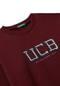 Felpa borgogna con collo a girocollo a coste, caratterizzata da un grande logo "UCB" in blu navy e bianco, con la scritta "ESTABLISHED 1965" stampata sotto.