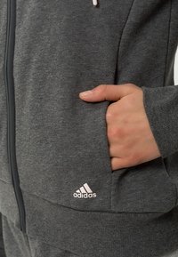 Main droite placée dans la poche d'un sweat à capuche Adidas gris foncé avec une fermeture éclair visible et un logo dans la partie basse de la poche.