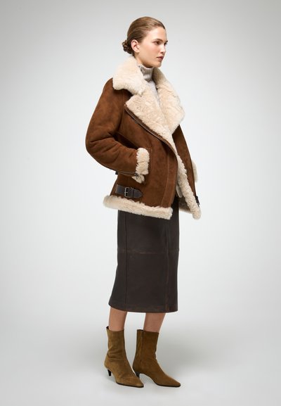 Brun shearlingjakke med en bred cremekrave, sølvhardware og sidebuckle. Modellen har en mørk læderskirt og grønne ankelstøvler på.