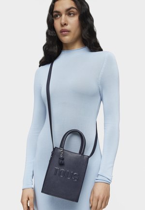 Bolso de piel azul marino con un diseño estructurado, que cuenta con un asa superior y una correa ajustable. En la parte frontal tiene un logo grabado de "TOUS".