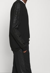 Černá mikina s dlouhými rukávy, se strukturálními černými panely na každém rukávu. Elastické manžety a volný střih. Jemný branding.