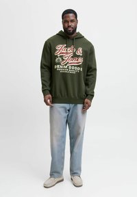 Olivengrøn hættetrøje med hvid og rød grafisk tekst, parret med lyseblå lige ben jeans og beige sko. Simpel design, afslappet pasform.