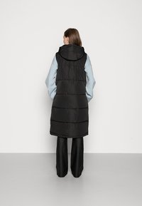 Gilet matelassé noir avec capuche, présentant un design matelassé et une longueur longue. Porté par-dessus un haut bleu clair, accompagné de pantalons noirs.