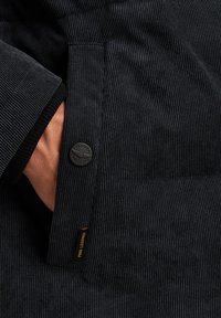 Le tissu en velours côtelé noir présente une poche avec un bouton noir rond et une petite étiquette de marque en or. La texture montre des lignes verticales distinctives.