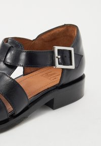 Black leather sandal featuring a wide strap design, adjustable ankle strap with silver buckle, and a low block heel. Interior has a brown finish.