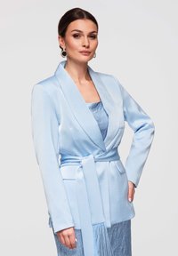 Femme portant un blazer bleu clair ceinturé sur une robe bleu clair texturée, avec des boucles d'oreilles en anneaux, posant devant un fond gris uni.