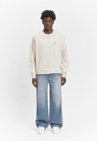 Overdimensioneret cremet sweater med ribbet tekstur, udstyret med et logo. Kombineret med løst siddende, blege blå jeans og hvide sneakers.