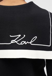 Îmbrăcăminte neagră tricotată cu margini albe și broderie cu litere cursive "Karl" pe partea superioară a spatelui, purtată de o persoană cu fața spre spate.