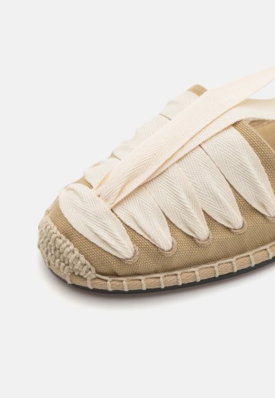Espadrille beige con parte superior de lona tejida y acentos crema. Presenta correas de tela gruesa y una suela de yute trenzado. Superficie texturizada.