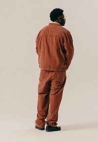 Muž s okuliarmi a náušnicou, ktorý má na sebe zladenú hnedú kockovanú bundu a nohavice, stojí bokom proti jednolitému pozadiu.