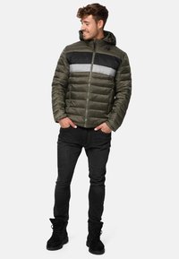 Grøn polstret jakke med en sort og grå vandret stribe, hætte, lynlås lukning og sidelommer; båret med sorte jeans og støvler.