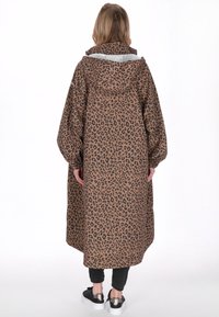 Barna leopárdmintás hosszú kabát kapucnival, hosszú ujjakkal és laza szabással. Fekete nadrág és tornacipő fölött viselve, puha tapintású anyagból.