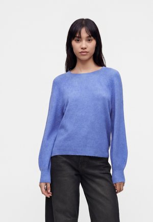 Ung kvinde iført en lyse lilla langærmet sweater og løse sorte bukser stående mod en ensfarvet hvid baggrund.