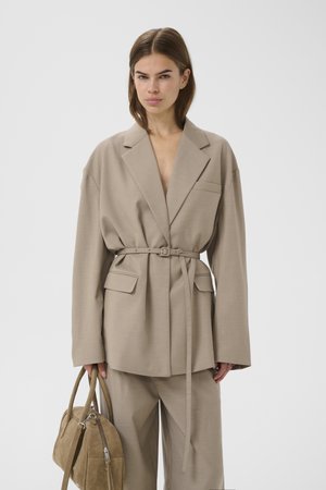 Kvinde iført oversized beige jakkesæt med bælte på blazeren og matchende bukser, der holder en tanfarvet tekstureret håndtaske mod en ensfarvet hvid baggrund.