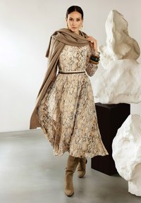 Beige Schlange-Muster Kleid mit ausgestelltem Rock, tailliertem Oberteil und einem braunen Gürtel. Das Model trägt einen braunen Schal und kniehohe Wildlederstiefel.