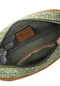 Interior de bolsa tejida verde con una etiqueta de cuero. Presenta un cierre con cremallera, un bolsillo y forro de tela oliva. Diseño simple y funcional.
