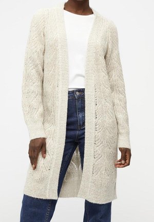 Personne portant un long cardigan crème ouvert en tricot, sur une chemise blanche et un jean bleu, debout sur un fond blanc uni.